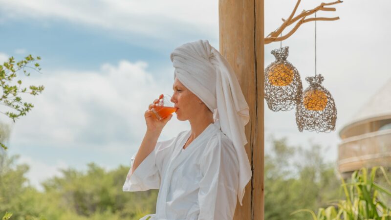 A person wearing a white robe and a towel wrapped around their head stands on a wooden deck, sipping a drink. Decorative lanterns hang from a tree branch beside them, evoking the ambiance of Soneva Exclusive Offers. The background features lush greenery and a partly cloudy sky.