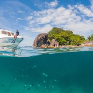 A person sits on the bow of a white boat anchored near a rocky shoreline with lush greenery under a blue sky with scattered clouds, savoring the tranquility. The water is clear, revealing small fish swimming near the surface—a true Soneva Exclusive Offers experience.