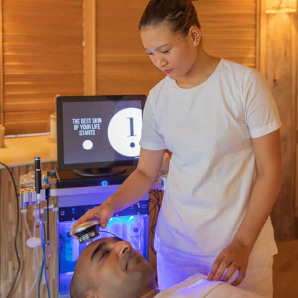A person receiving a facial treatment lies on a spa bed covered with a white sheet at a Maldives spa. A spa worker in a white uniform applies a device to the person's forehead. In the background, wellness equipment with a screen displays "THE BEST SKIN OF YOUR LIFE STARTS!