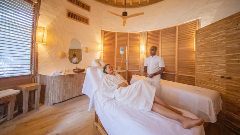 A woman lies on a treatment bed in a peaceful Maldives spa room with wooden decor, soft lighting, and a ceiling fan. She is getting an IV drip while a spa attendant in white attire stands beside her. The wellness room features another treatment bed and a vanity with a round mirror.