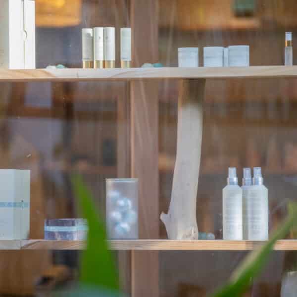 A wooden shelf displaying various skincare and beauty products, including bottles, jars, and boxes of creams, lotions, and serums reminiscent of a Maldives spa. Some of the products are placed on glass holders. Green plants partially frame the view of the shelf, adding to the wellness vibe.