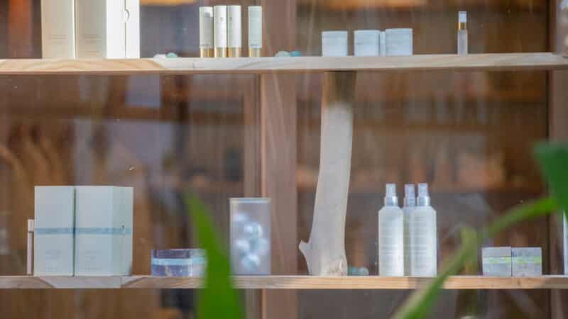 A wooden shelf displaying various skincare and beauty products, including bottles, jars, and boxes of creams, lotions, and serums reminiscent of a Maldives spa. Some of the products are placed on glass holders. Green plants partially frame the view of the shelf, adding to the wellness vibe.