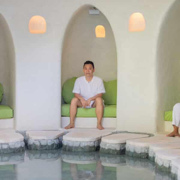 Three people dressed in white are sitting in an arched alcove area, with green cushions behind them. They are positioned above a shallow pool of water with stepping stones leading up to them. The setting is tranquil, reminiscent of a Maldives spa, featuring soft, warm lighting from the ceiling.