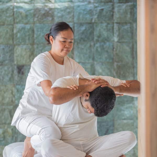 A massage therapist in white attire is performing a stretching technique on a seated client at a serene Maldives spa. The client, also dressed in white, sits cross-legged on a massage table as the therapist applies pressure on their back against the tranquil tiled background, promoting wellness and relaxation.