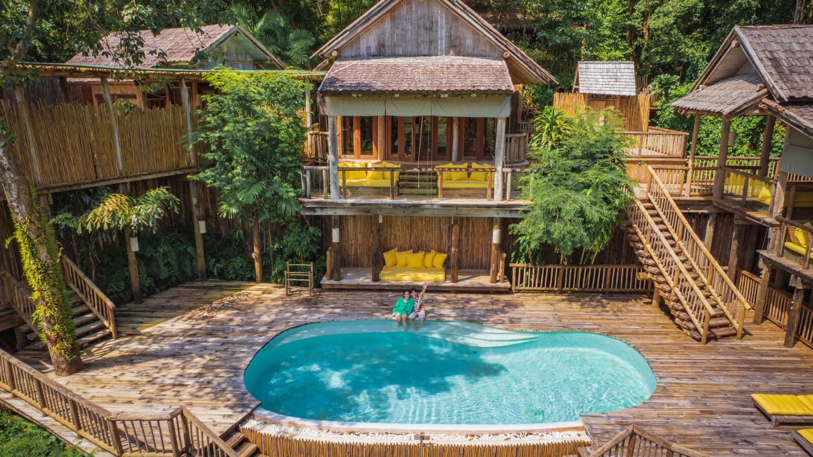 A tropical wooden villa surrounded by lush greenery, featuring a central swimming pool with two people lounging near the water's edge. Wooden staircases and railings lead to various parts of the villa, and vibrant yellow furniture accents the rustic setting. Discover more with Soneva Exclusive Offers.