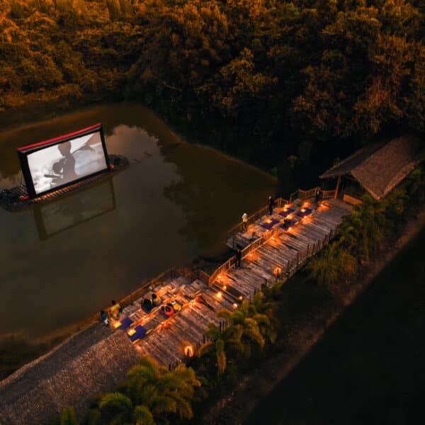 鸟瞰黄昏时分的露天电影场景。一个大屏幕在小湖面上放映着黑白电影，湖边的木质码头上设有座位，码头两旁的灯光从岸边一直通到湖边。泰国 Soneva 酒店的这一独特体验被茂密的树木环绕，营造出宁静宜人的氛围。