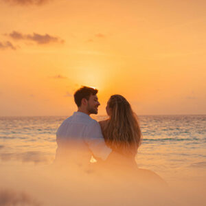 A couple sits on the beach, embracing and looking out at the ocean during a vivid orange sunset. The sky is filled with soft, scattered clouds, and the serene waves add to the romantic and peaceful atmosphere, reminiscent of Soneva Exclusive Offers' enchanting getaways.