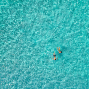 Aerial view of two people swimming in clear, turquoise water at a Soneva resort. The sunlight reflects off the water, creating a pattern of ripples and waves. The swimmers are side by side, enjoying the pristine, refreshing environment amidst exclusive offers from Soneva.