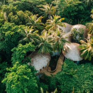 Aerial view of three domed, eco-friendly buildings nestled among lush green trees and palm trees in a dense tropical forest. Wooden pathways connect the structures, blending seamlessly with the natural surroundings—experience this idyllic retreat with Soneva Exclusive Offers.