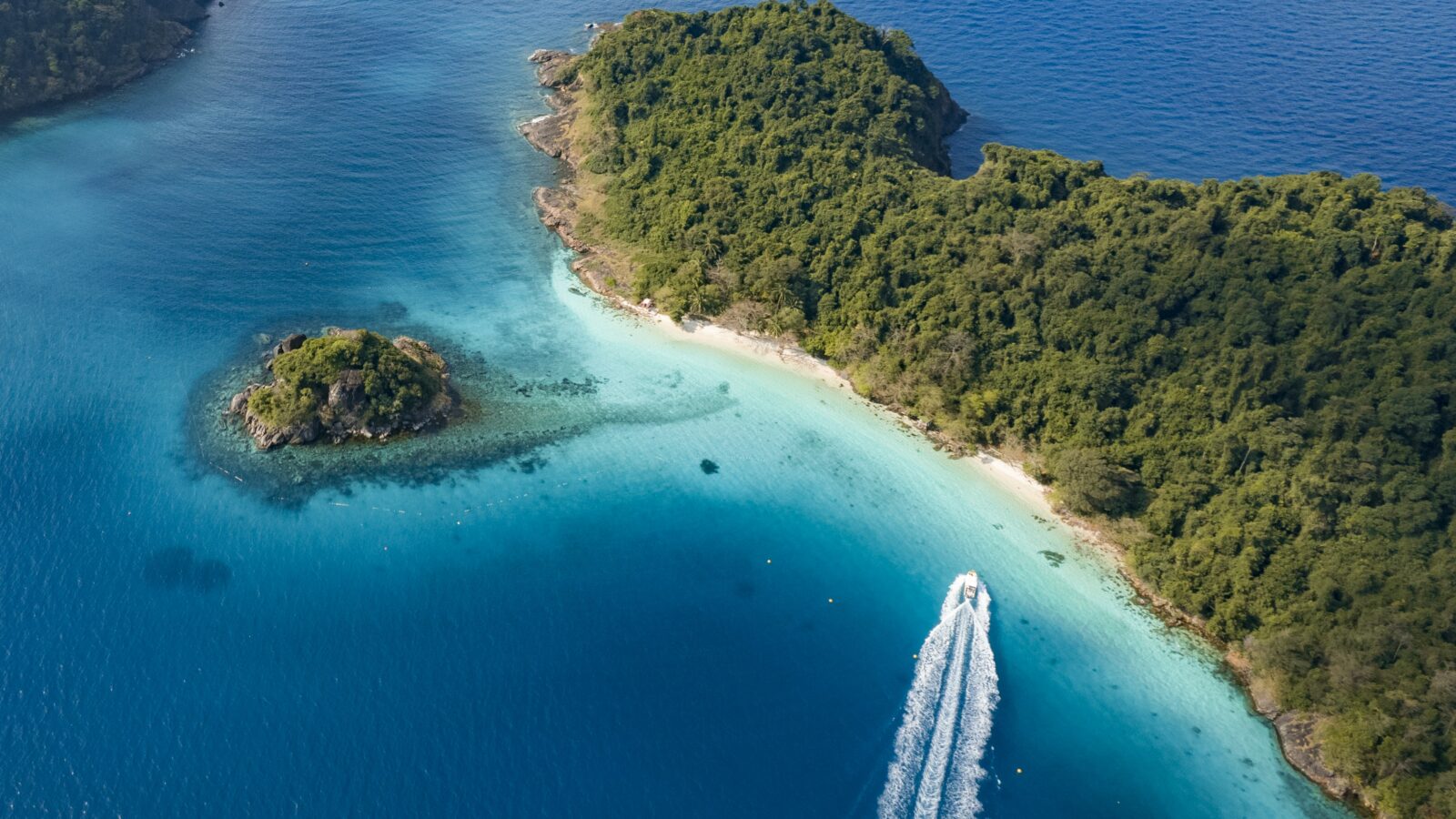 Aerial view of a speedboat leaving a white wake as it zips through clear blue waters near the tropical coastline of Soneva Kiri Resort. The shoreline is lined with lush, green vegetation, and a small, forested island sits nearby.