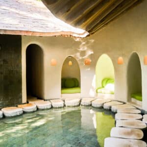 A serene indoor spa features a shallow pool with stone stepping paths leading to alcoves with cushioned seating. Sunlight streams through small rounded windows, softly illuminating the Maldive Spa Wellness-inspired space that exudes a tranquil, earthy ambiance with natural textures.