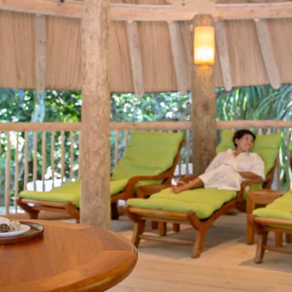 Two individuals relax on green lounge chairs in a serene outdoor setting surrounded by lush foliage, reminiscent of a Maldives spa. Wooden beams and a thatched roof give the space a rustic feel. A wooden table in the foreground holds drinks and snacks, adding to the tranquil, wellness-focused ambiance.