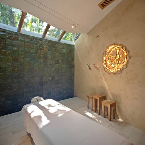 A serene Maldives spa wellness room featuring a massage table draped in white linens, rustic wooden stools, soft lighting, and wall art designed to resemble glowing petals. Skylights in the ceiling let in natural light, enhancing the peaceful atmosphere.