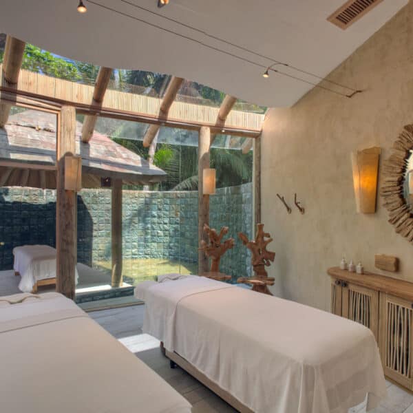 A serene spa treatment room featuring two massage tables with white linens. The room has a rustic design with wooden elements, soft lighting from natural sunlight, and earthy-toned walls. A large mirror with a wooden frame and nature decor evoke a Maldives spa wellness atmosphere.