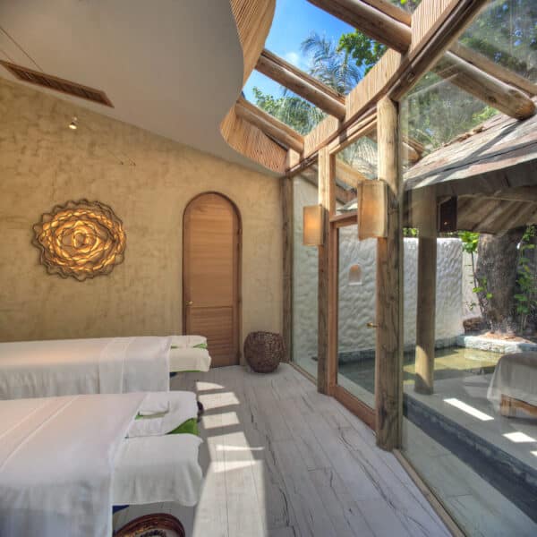 A serene spa room with two massage tables covered in white linens, positioned near a large window. The space is filled with natural light, featuring rustic wooden elements, textured walls, a decorative wall piece, and views of lush greenery reminiscent of Maldives spa wellness.