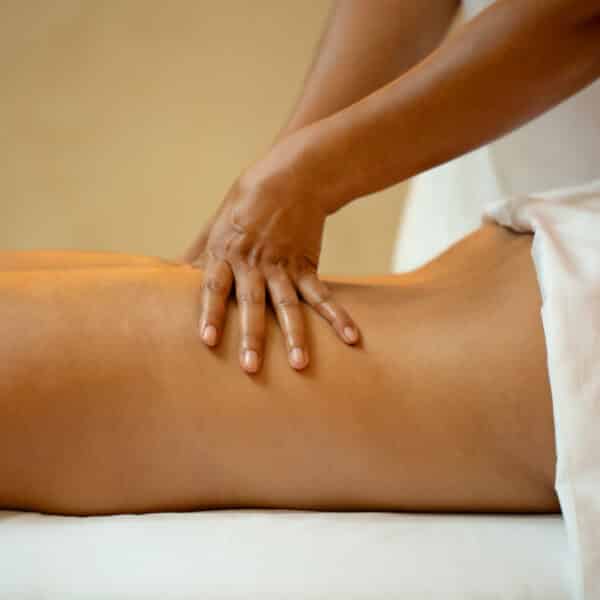 A person lies face down on a massage table, partially covered with a white towel. At this serene Maldives spa, the therapist's hands are shown applying pressure to the person's mid-back area. The background is softly blurred.
