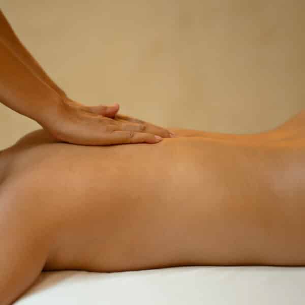 A person's back being massaged with both hands visible applying pressure. The individual is lying face down on a massage table with a white sheet draped over the lower part of their body. The background, reminiscent of a Maldives spa, is softly lit and blurred, enhancing the sense of wellness.