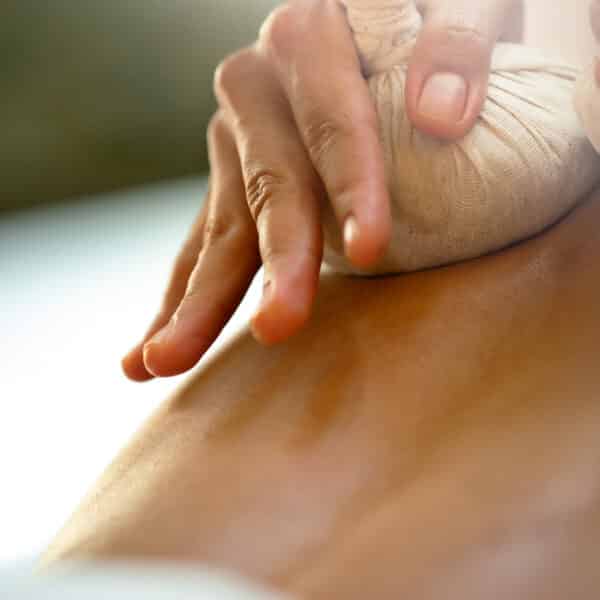 Close-up of a person receiving a massage with a herbal compress at a Maldives spa. A hand is pressing a small cloth-wrapped bundle against the person's back, creating a therapeutic and relaxing atmosphere. The background is softly blurred, emphasizing the focus on this wellness treatment.