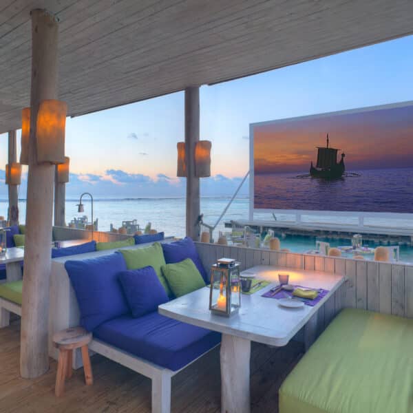 A seaside restaurant with wooden furniture and vibrant green and blue cushions. Tables are set with candles and place settings. The view includes a sea with a large screen displaying a sunset scene of a boat on the water. Lanterns hang from wooden pillars, offering an exclusive Maldives experience reminiscent of Soneva&#039;s luxury.