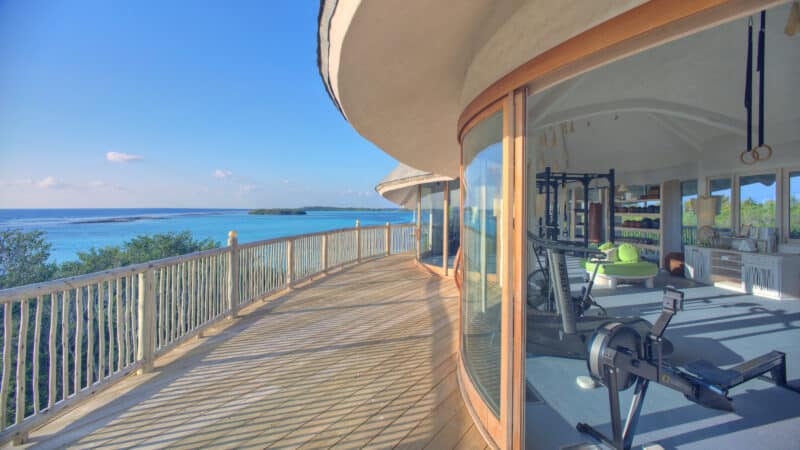 A sunlit balcony overlooks a serene ocean and greenery. The adjoining room, enclosed with curving glass walls, is equipped as a modern gym with various exercise machines, offering a Maldive Spa Wellness experience.