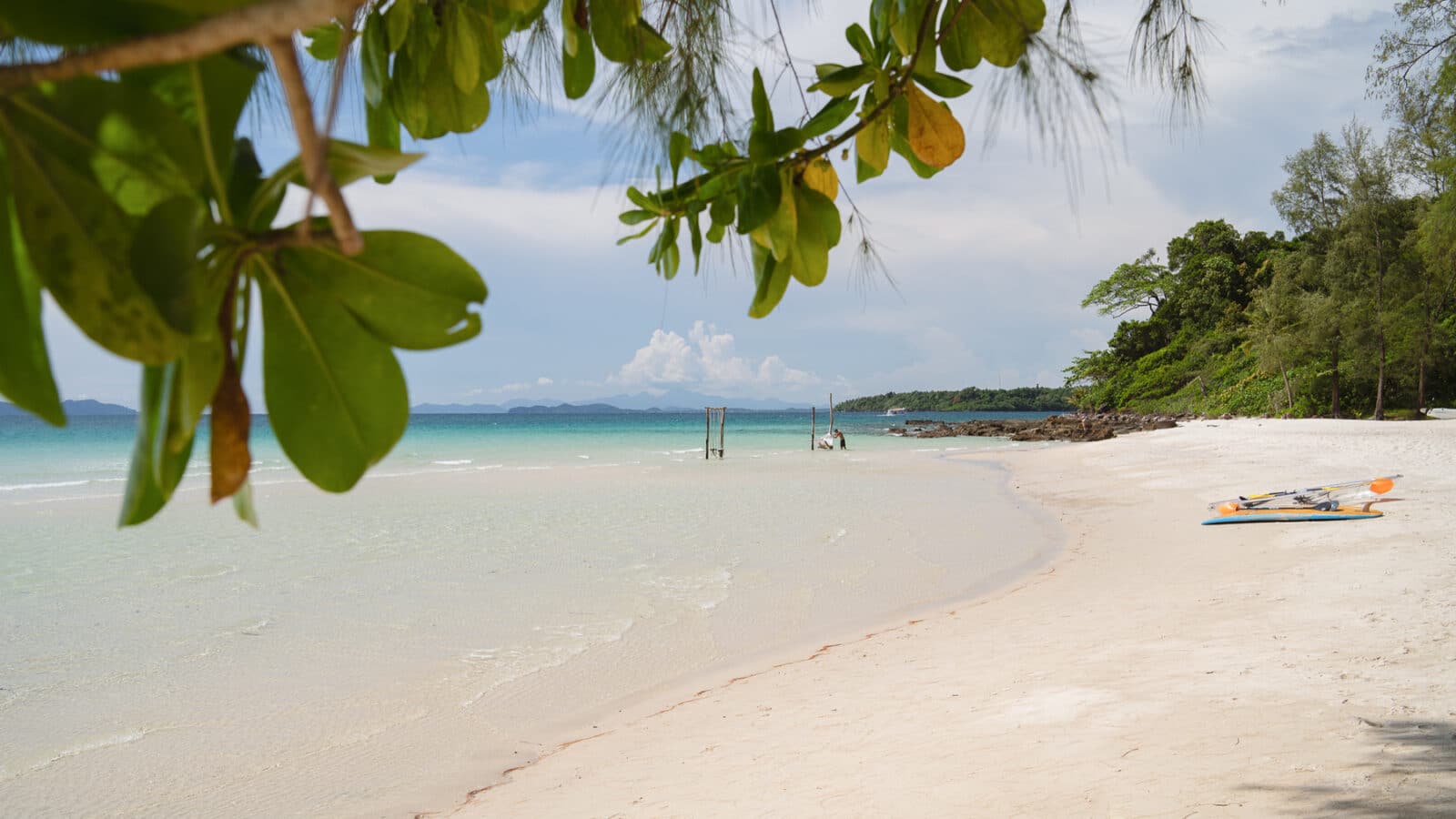 A serene beach scene with clear turquoise waters, white sand, and lush greenery. Kayaks rest on the sand while a small wooden structure stands in the shallow water. Overhanging tree branches with green leaves frame the top of the image, offering a Thailand exclusive experience reminiscent of Soneva’s tranquility.