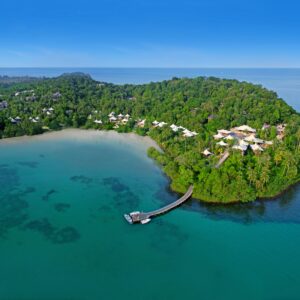 An aerial view of a tropical island with lush greenery surrounded by clear turquoise waters. A curved pier extends from a cluster of buildings in the forested area out into the sea, connecting the island to a small dock amidst the tranquil blue waters, reminiscent of Soneva Exclusive Offers.