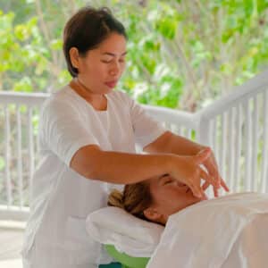 A massage therapist in white attire is performing a facial massage on a client lying on a massage table covered with a white sheet. The background features lush green foliage, reminiscent of the tranquil ambiance at a Maldives spa, suggesting an exquisite outdoor or wellness setting.