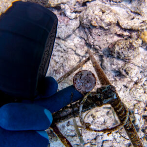 A gloved hand holds a small, aged coin with the number "15" on it, discovered among rusted metal debris underwater near the Maldives. The seabed is visible in the background, characterized by rocky, coral-like formations.