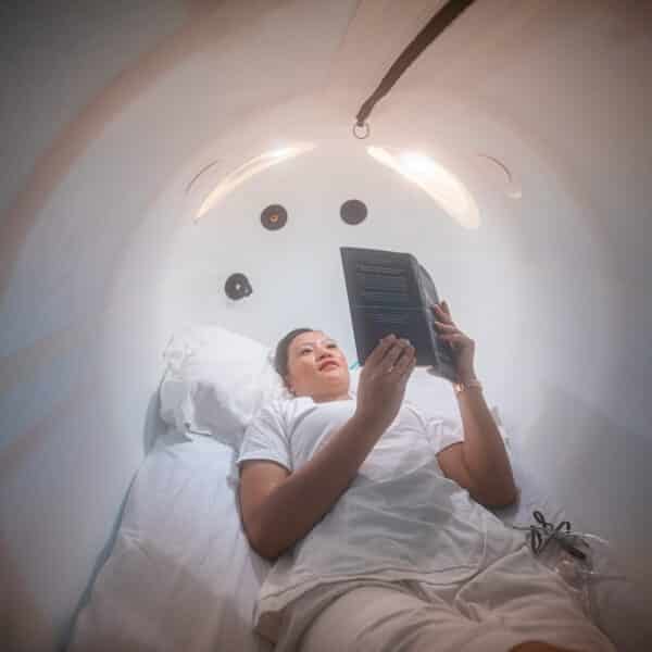 A woman wearing a white shirt lies on a bed inside a circular hyperbaric oxygen therapy chamber at Soneva Soul, reading a book. The chamber, reminiscent of the Maldives spa wellness, features small windows and medical cables. She appears relaxed and focused on her reading.