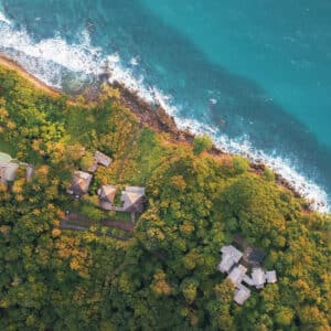 Aerial view of a lush, green coastal area with dense vegetation and scattered houses. The coastline features rocky edges meeting the ocean, with waves crashing against the shore. Adjustments in terrain suggest a slightly elevated forested area leading to the water, reminiscent of Soneva Exclusive Offers.