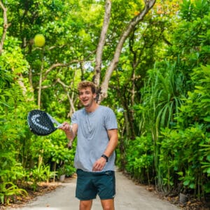 A man in a gray t-shirt and dark shorts is playing tennis in the middle of a lush, green forest as part of the exclusive Soneva experience. He is holding a racket and hitting a tennis ball amid dense greenery and tall trees, evoking the tranquility of the Maldives.