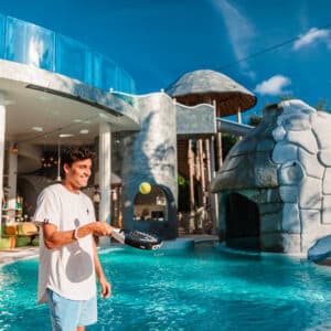 A person in casual attire stands by an elaborate poolside resort area, enjoying Soneva Exclusive Offers while playing with a paddle and ball. The resort features a water slide, colorful loungers, and a futuristic dome-like structure under the clear blue sky.