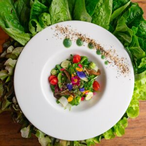 一个白色盘子，上面摆放着五颜六色的美食沙拉，有混合蔬菜、樱桃番茄、食用花和其他蔬菜。盘子上点缀着绿色酱滴和调味料，周围是木质表面上的新鲜生菜叶。体验 Soneva 独家优惠，品尝这样的美食佳肴。