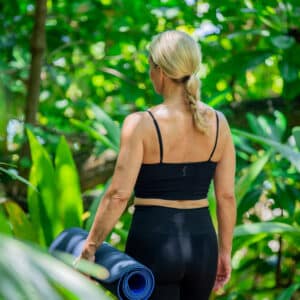 A woman with blonde hair is wearing a black sports bra and leggings. She stands outdoors in a lush, green forest setting, holding a rolled-up yoga mat. Her back faces the camera as she gazes into the distance, embodying the tranquility of Soneva Exclusive Offers.