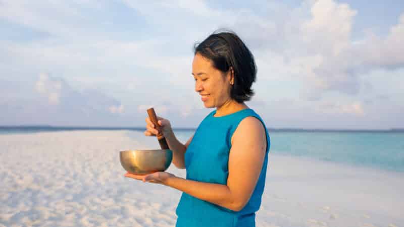 A woman stands on a sandy beach in the Maldives, holding a singing bowl and mallet. She wears a sleeveless blue top and is smiling while striking the bowl. The ocean and a partially cloudy sky are visible in the background, evoking a serene sense of spa wellness.