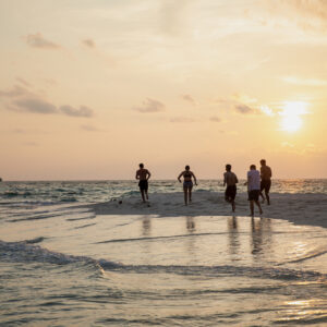 A group of people running towards the horizon on a beach during sunset, embodying Maldives wellness. The ocean waves gently lap at the shore, and the sky is painted in warm hues of orange and yellow.
