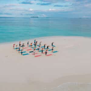 A group of people practices yoga on a pristine, sandy beach at a Maldives spa. Vibrant blue and red yoga mats are spread out in neat rows on the white sand. The calm, turquoise ocean and a partly cloudy sky create a serene and picturesque background, perfect for wellness activities.