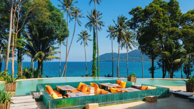 An outdoor seating area with vibrant yellow and orange cushions is situated in the middle of a shallow pool. Tall palm trees and lush greenery surround the space, with a clear blue ocean and mountainous island visible in the background under a bright sky—truly an exclusive Soneva experience in Thailand.