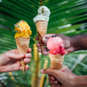 Three hands holding ice cream cones against a backdrop of lush green palm leaves. Each cone boasts a different flavor: yellow with sprinkles, green, and pink. The scene suggests a tropical, summery atmosphere—perfect for enjoying Soneva Exclusive Offers tailored for your dream getaway.