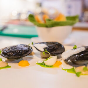 A gourmet dish featuring three black-colored ravioli served with dollops of white and yellow sauces and green garnish, all artistically arranged on a white plate. The presentation evokes the elegance of a Maldives Luxury Yacht Holiday, with a blurred background that includes a bowl of greens.