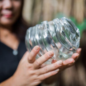 A person holding a broken glass jar with both hands, sharp, jagged edges where it has shattered. The background is blurred, drawing attention to the jar and the person's hands with light-colored nail polish—a stark contrast to the serene beauty of a Maldives exclusive experience at Soneva.