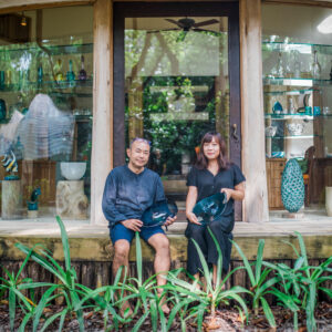 A man and a woman sit in front of a wooden-framed glass storefront displaying various pottery pieces, reminiscent of a Maldives Exclusive Experience. Both are holding black ceramic creations and appear relaxed. Green plants grow in the foreground, adding a natural touch to the scene.