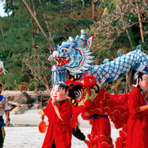 Performers in colorful costumes, including vibrant red robes and large masks, participate in a traditional dragon dance on a sandy beach at Soneva. Celebrating Chinese New Year in the Maldives, a blue dragon puppet weaves through the scene, backed by lush greenery and trees.
