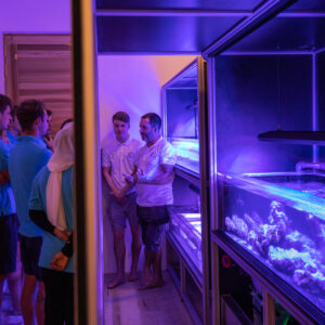 A group of people stands in a dimly lit room with purple lighting, listening to a man gesturing towards a large aquarium. The setting, reminiscent of a laboratory or research facility, is part of an event organized by the Soneva Charity Foundation Maldives.