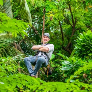 A person wearing a helmet and harness glides through a lush, green forest canopy on a zipline, smiling. The background is filled with vibrant leaves and dense foliage, offering an adventure reminiscent of a Soneva Exclusive Experience in the Maldives.