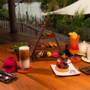 A wooden table set with an array of desserts and beverages, including tarts, macarons, a layered coffee drink, a fruity cocktail, and a teapot with cups. The setting is outdoor with lush greenery and a touch of water in the background, creating a tropical ambiance reminiscent of a Thailand Exclusive Experience at Soneva.