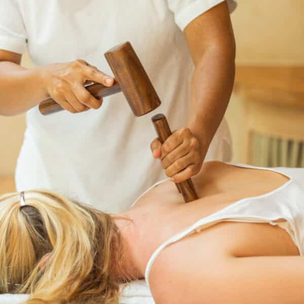 A person receiving a therapeutic massage lies face-down on a massage table at a Maldives spa. The massage therapist gently uses wooden mallets in a technique that involves tapping on the person's back to provide muscle relaxation. The individual, with blonde hair tied in a ponytail, appears calm and relaxed amidst the wellness ambiance.
