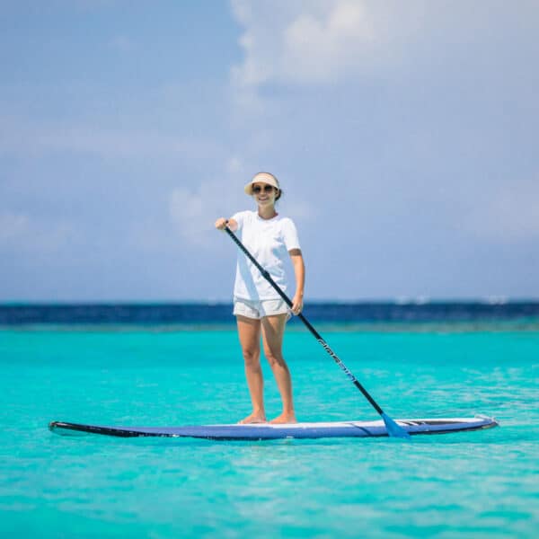 A person wearing a white shirt, white shorts, and a hat is standing on a paddleboard in the clear, turquoise water of a Soneva Exclusive Experience Maldives. They are holding a paddle and smiling. The sky is bright with some clouds, and the horizon is visible in the background.