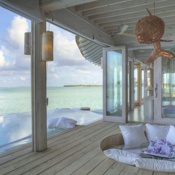 Spacious, open-air room with panoramic ocean views. A sunken seating area with white cushions is in the center, surrounded by wood flooring. Doors lead to a bedroom, and to the left, there's an infinity pool merging with the sea. Décor includes beach-themed elements perfect for Maldives villa ownership.