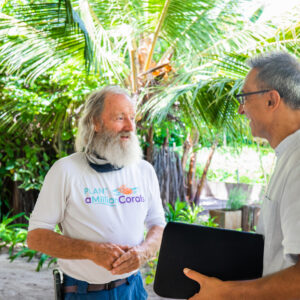 Two older men stand outdoors in a tropical setting with lush greenery and palm trees. One, with a long white beard, is wearing a "Plant a Million Corals" shirt and a neck gaiter, while the other, representing the Soneva Charity Foundation in Maldives, holds a black folder and smiles engagingly.